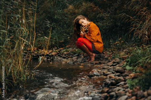 Fototapeta Naklejka Na Ścianę i Meble -  a girl with long hair and an orange sweater and red tights holds a dog in her arms. In the open air by the stream.