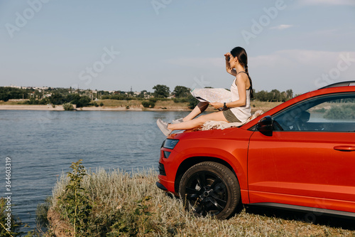 Young woman sitting on the car hood, holding a map, on a coast by a lake. Concept of tourism. © Bostan Natalia