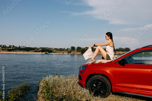 Young woman lying on car hood  holding a map  looking towards a lake. Road trip concept.