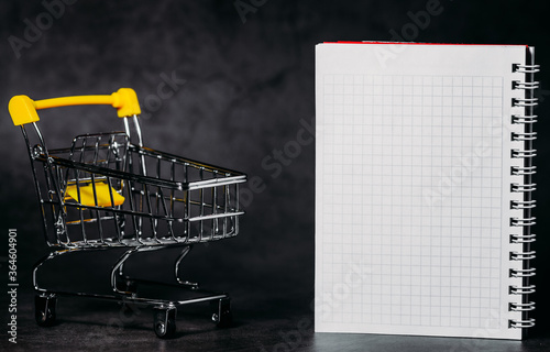 Shopping trolley and blank notebook on a dark background. Conceptual, copy space.