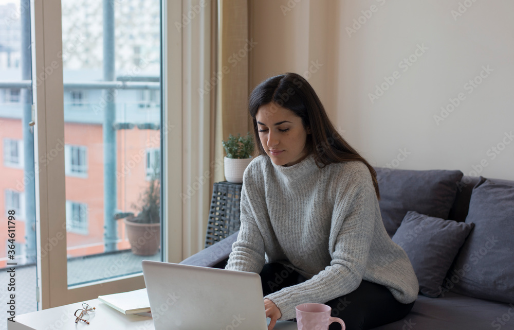Young woman working on a laptop by the window at home in Birmingham, Uk. Young woman working on a laptop by the window at home in Birmingham, Uk.