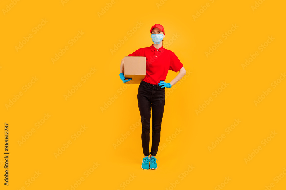 Full length, young female courier dressed in a Red cap and t-shirt, uniform, medical protective mask and gloves, holds cardboard boxes and shows a gesture of strength against a yellow background Full length, young female courier dressed in a Red cap and t-shirt, uniform, medical protective mask and gloves, holds cardboard boxes and shows a gesture of strength against a yellow background
