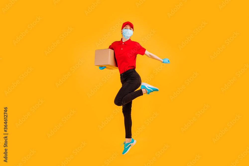 A cheerful, excited, Bouncing female courier in a red Cap and t-shirt, uniform, medical protective mask and gloves, with boxes in her hands, on a yellow background. coronavirus