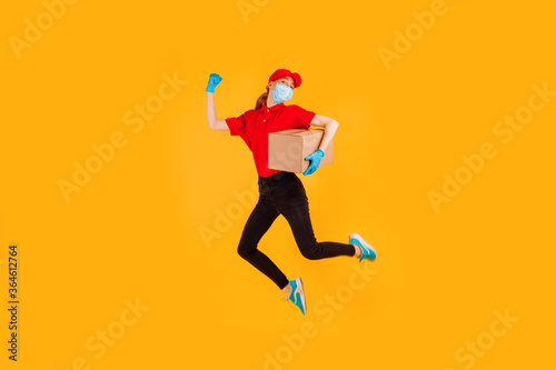 A cheerful, excited, Bouncing female courier in a red Cap and t-shirt, uniform, medical protective mask and gloves, with boxes in her hands, on a yellow background. Delivery service © Shopping King Louie