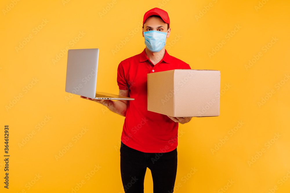 male courier, in a red uniform, wearing a medical mask and gloves, holds a cardboard box and a laptop, on a yellow background. Delivery service, online shopping, quarantine, coronavirus male courier, in a red uniform, wearing a medical mask and gloves, holds a cardboard box and a laptop, on a yellow background. Delivery service, online shopping, quarantine, coronavirus
