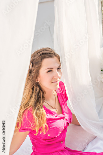 A cute blonde girl in a crimson magenta long dress sits on a bed with white sheets. The iron sofa is decorated with Chiffon canopies at the top, white wisteria, flowers. Gentle, romantic morning 