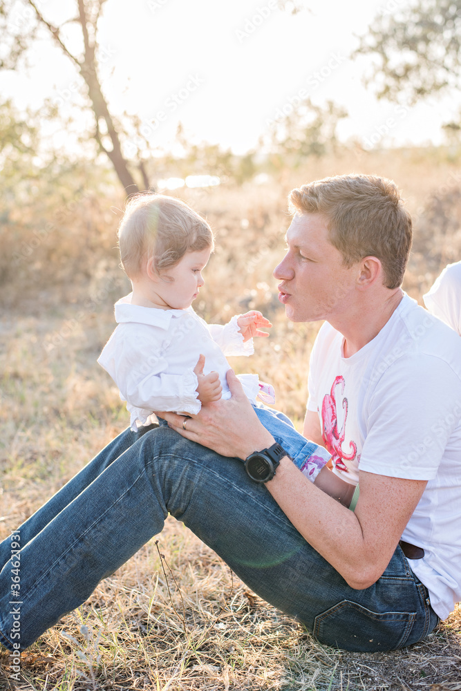 Fototapeta premium A young red-haired man holds a small beautiful daughter in his arms. She rolls it on her shoulders, plays, shakes, carries on hands. The baby laughs in her two teeth. Parenthood concept, child care
