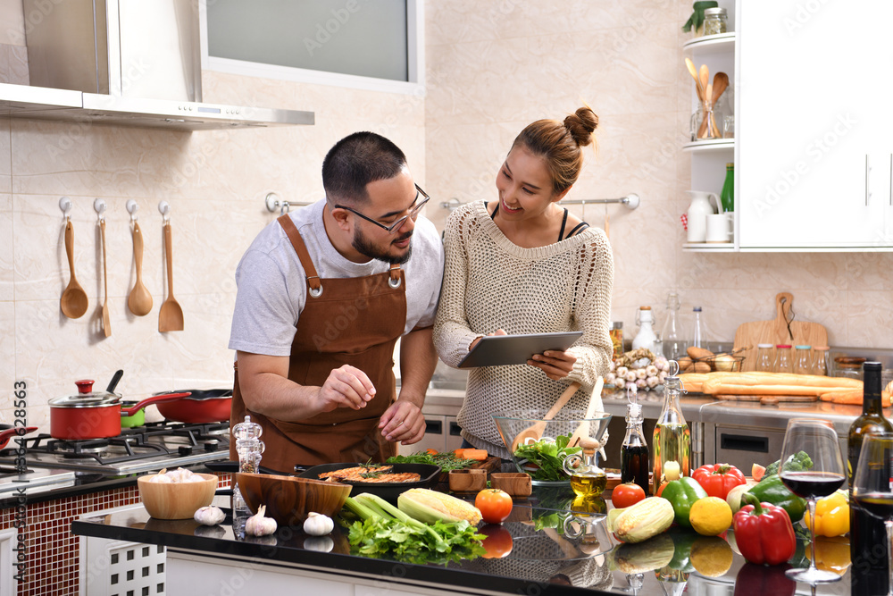 Loving young Asian couple using digital tablet and cooking in kitchen making healthy food together feeling fun - obrazy, fototapety, plakaty 