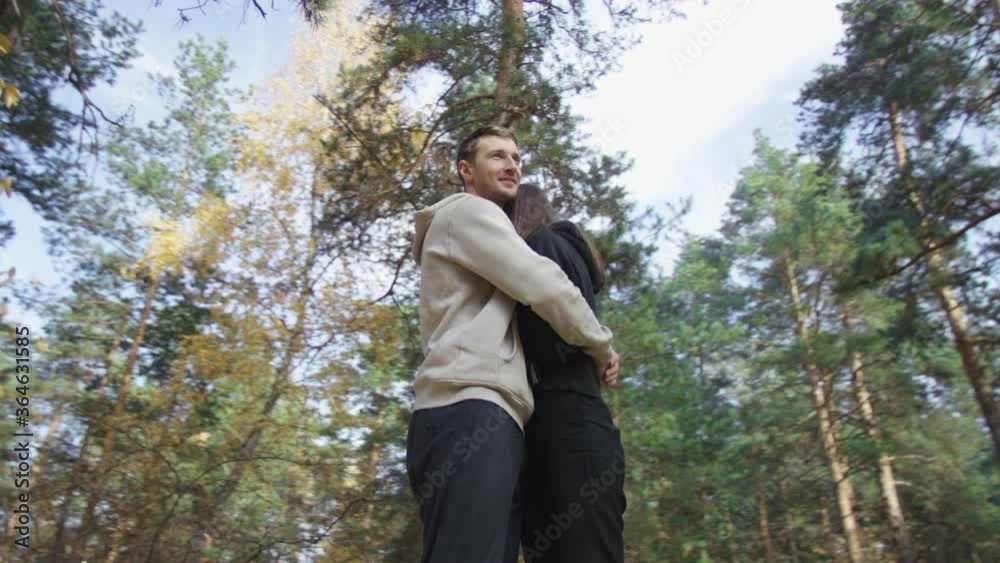Handsome man soothingly embosoms attractive female. Couple standing in brightly lit autumn forest. Low angle 4K 360 degree tracking arc shot.