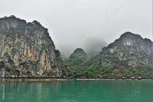 Islands in Halong Bay in the fog. Calm emerald water and a narrow strip of sand on the shore. Bizarre rocky islands hide in a foggy haze. UNESCO Heritage. Vietnam. 