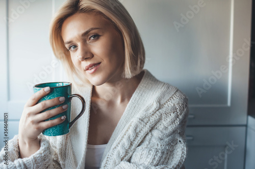 Smiling lovely woman with blonde hair drinking a cup of tea in the kitchen wearing a knitted sweater