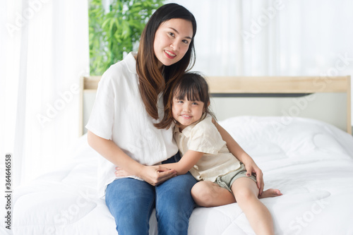 Lovely Asian mother and little daughter hug together, smile and feeling happy on the bed in a cozy house, look at camera, family concept