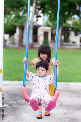 Younger brother sat on swing and his sister was helping to swing the blue swing. Activities and mutual care in family. Children playing in playground. Background is bright green lawn. Summer or spring