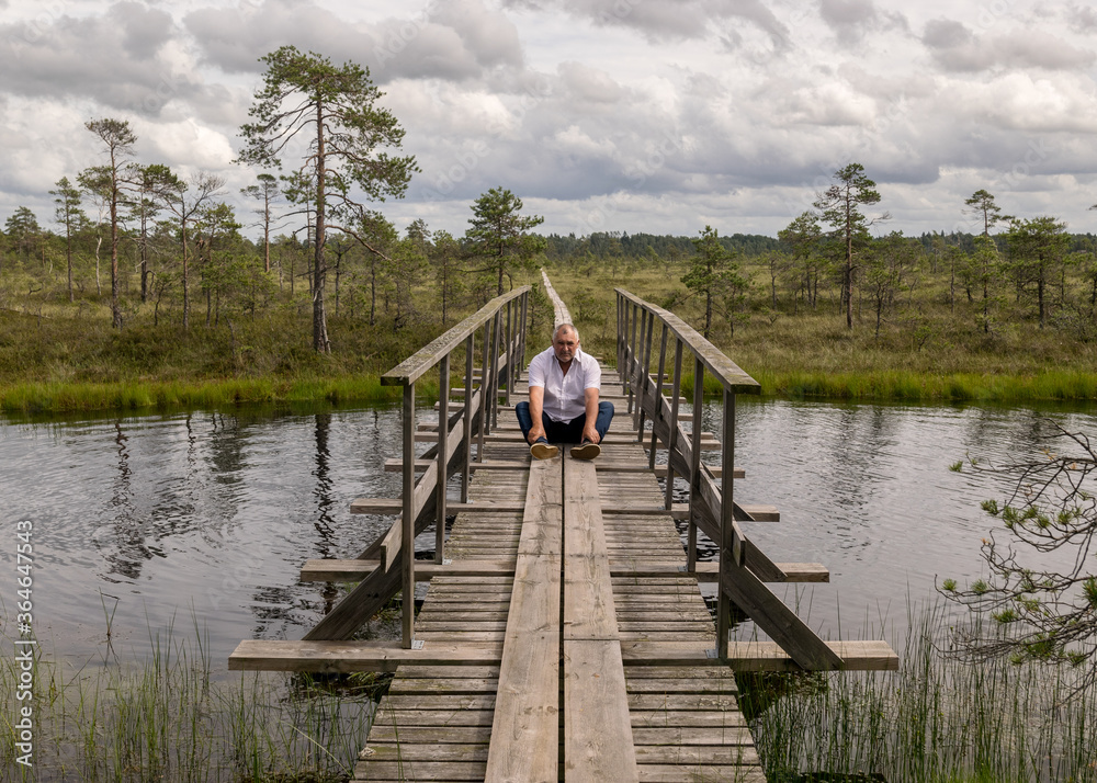 the summer swamp. a man in a white shirt sits on a wooden bridge. bog pond. bog background and vegetation the summer swamp. a man in a white shirt sits on a wooden bridge. bog pond. bog background and vegetation