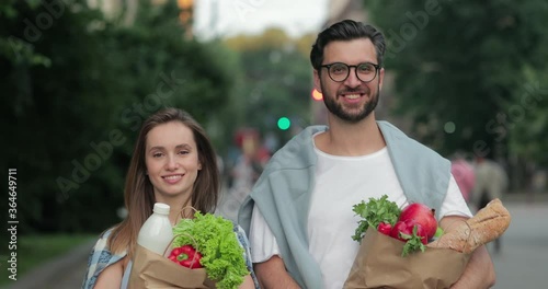 Crop view of young cheerful couple walking at street and holding paper bags with food. Good looking man and woman looking to camera and smiling while carrying their shopping photo