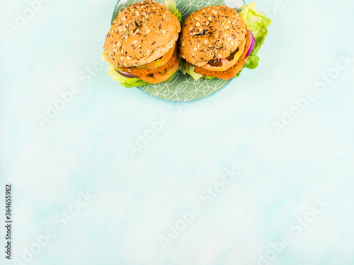 Vegan burgers with soy and vegetable patties, onion, tomatoes and lettuce with spelt and seeds buns on green dish over turquoise background. Copy space, flat lay