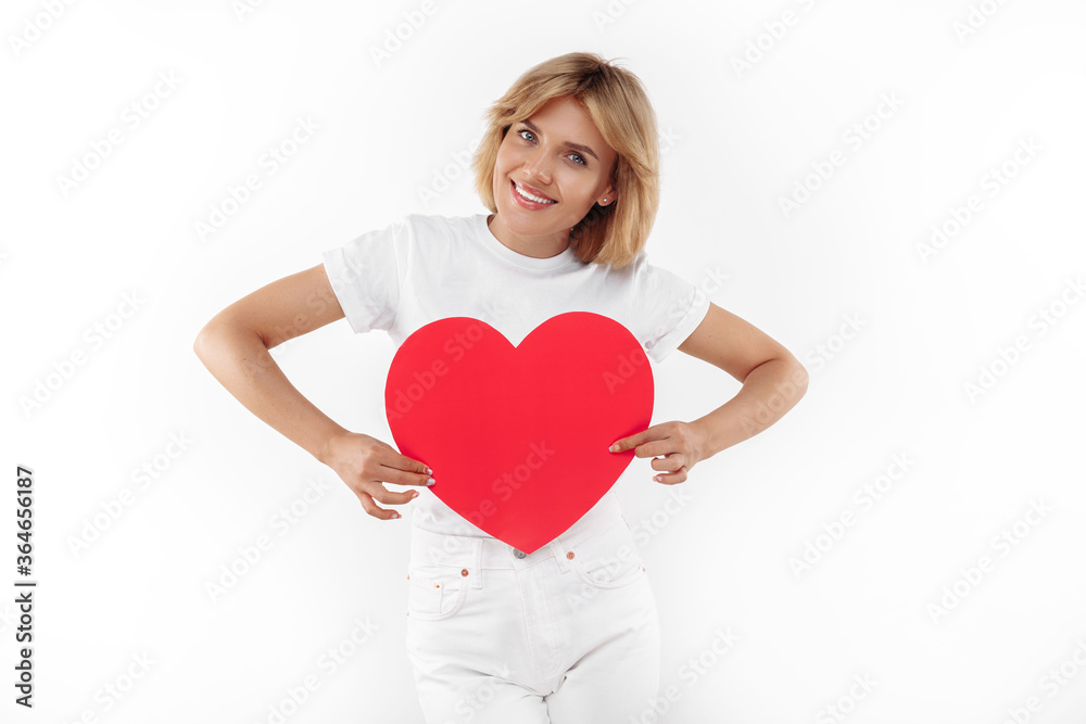 Positive young blonde woman in casual white outfit holding paper heart over white background. Positive young blonde woman in casual white outfit holding paper heart over white background.