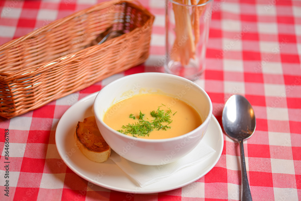 Pumpkin soup in a white bowl. Wooden table, red plaid tablecloth, blurred background. Dinner in restaurant. Pumpkin soup in a white bowl. Wooden table, red plaid tablecloth, blurred background. Dinner in restaurant.