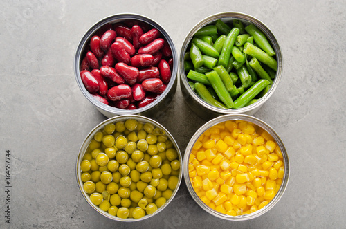 Flat lay view at canned kidney beans, green beans, peas and corn in opened tin cans on kitchen table. Non-perishable foods background