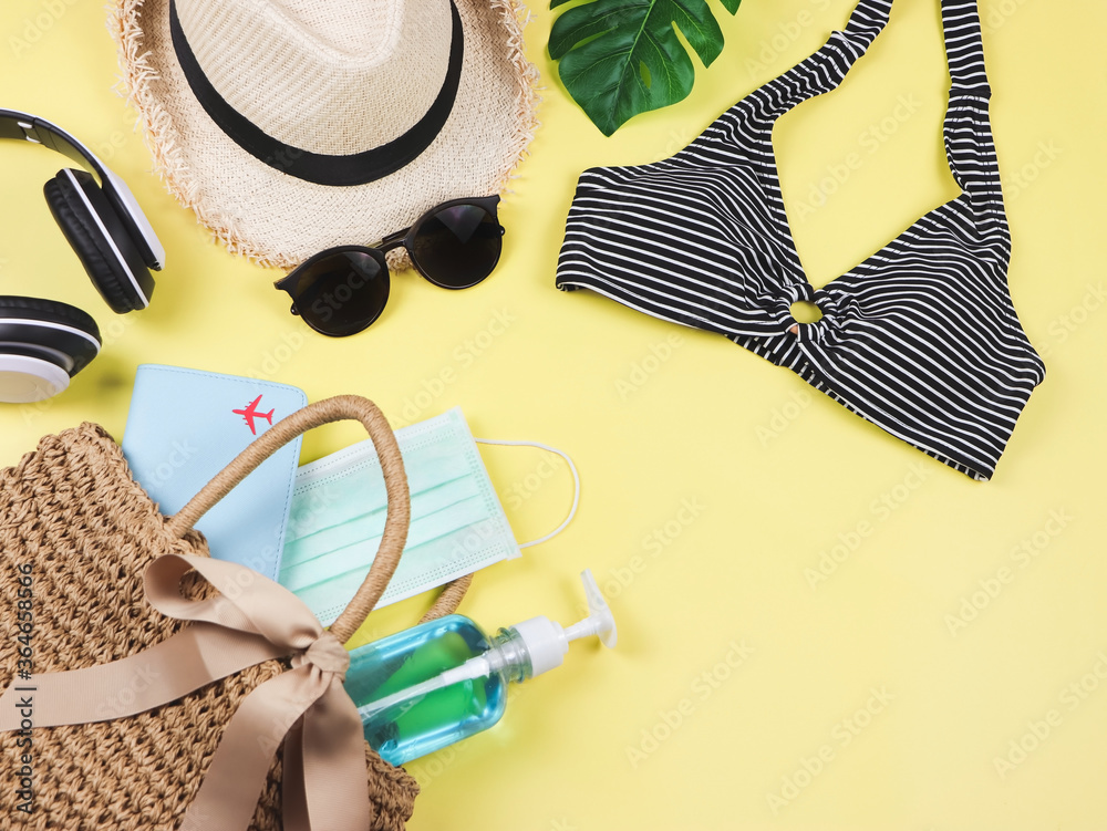 COVID-19 prevention , summer and new normal concept, top view of   bikini swim wear  and women's vacation accessories with medical mask and sanitizer gel  in woven bag on yellow background. - obrazy, fototapety, plakaty 