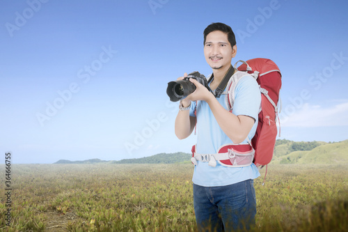 Asian man with a backpack holding the camera on the meadow field