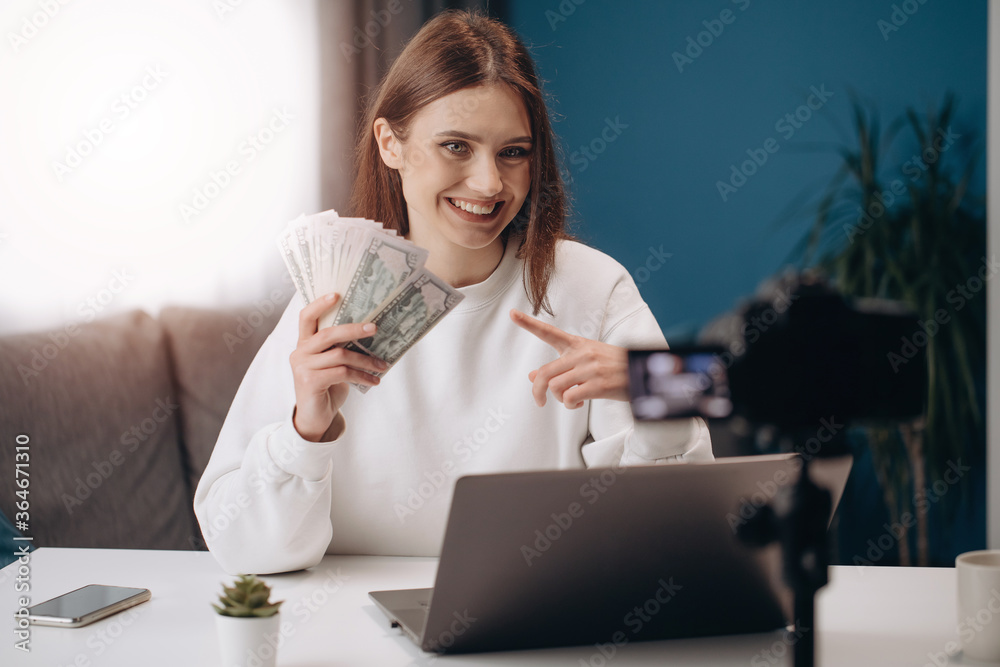 Smiling female blogger with brown hair sitting at table with dollars banknotes while recording video on camera. Young woman in white sweater teaching her followers how to earn money online. Smiling female blogger with brown hair sitting at table with dollars banknotes while recording video on camera. Young woman in white sweater teaching her followers how to earn money online.