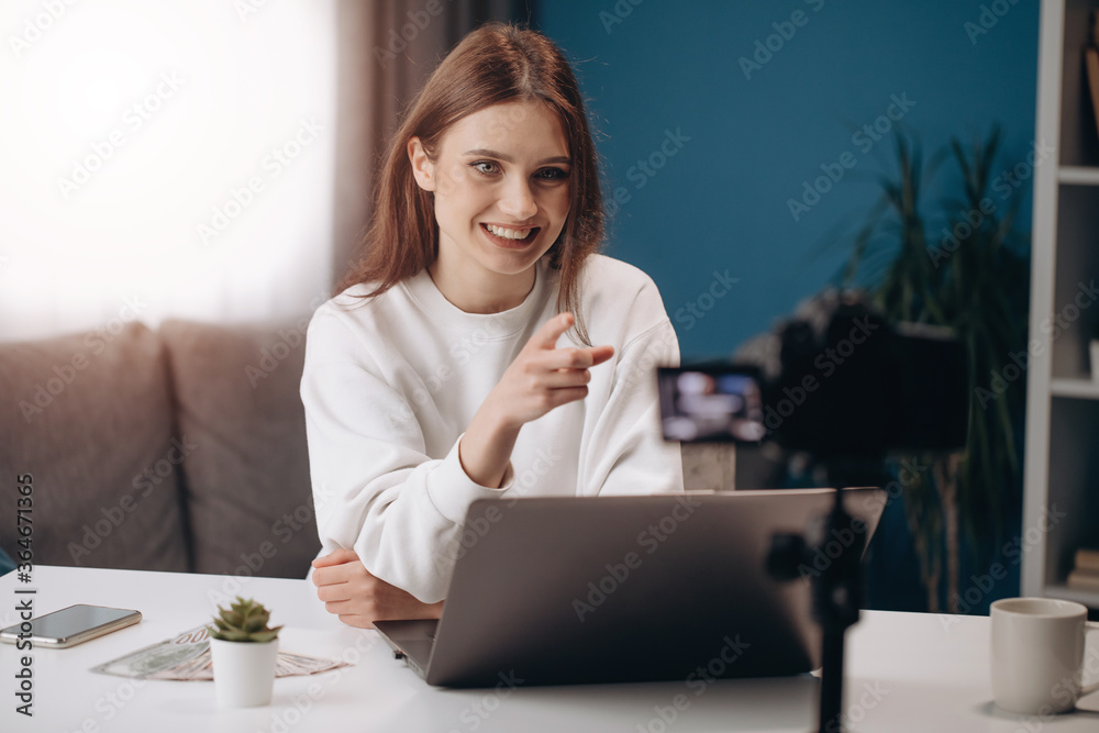 Beautiful woman in casual outfit sitting at table with opened laptop and money cash while filming video on digital camera. Concept of tutorials, blogging and social networks Beautiful woman in casual outfit sitting at table with opened laptop and money cash while filming video on digital camera. Concept of tutorials, blogging and social networks
