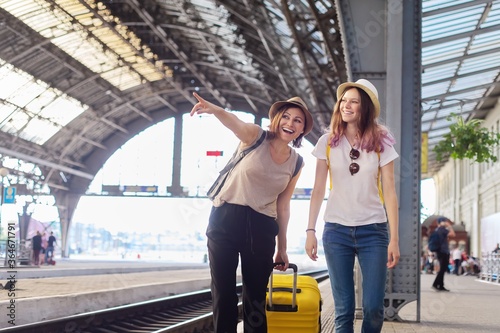 Happy mother and teenager daughter walking with suitcase at railway station