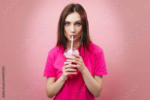 Emotional brunette female in casual t-shirt standing over pink background and drinking tasty cocktail with ice cubes. Happy woman refreshing during summer time.