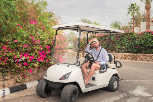 A man straightens his hat while sitting in a white golf cart. The guy is sitting in an electrocar next to flowers and palm trees. © Климов Максим