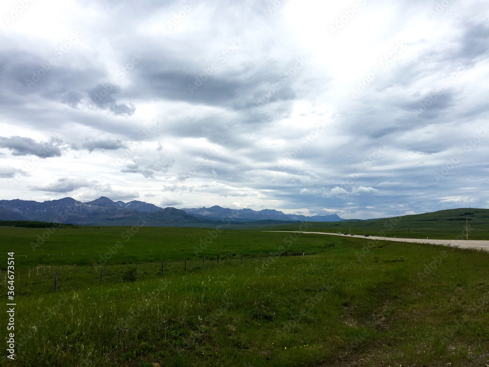 Dramatic clouds with beams of sun, range of mountains on horizon. Road trip to Rocky mountains in Alberta, Canada. No people on farm field, long road leading to mountains with few barely visible cars