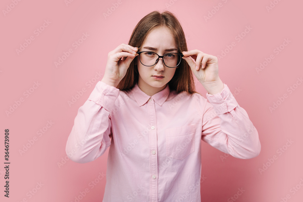Beautiful blonde girl in pink shirt holding hands on eyeglasses and looking at camera with suspicious glance. isolated over studio background. Concept of real human emotion. Beautiful blonde girl in pink shirt holding hands on eyeglasses and looking at camera with suspicious glance. isolated over studio background. Concept of real human emotion.