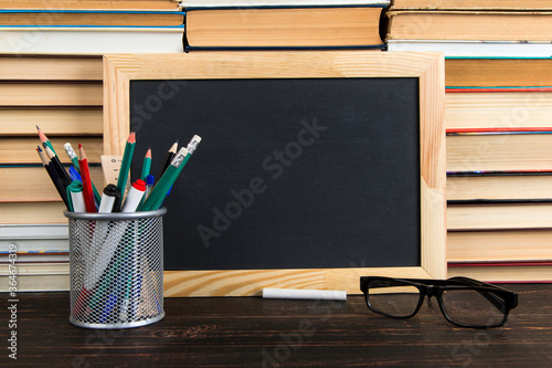 Chalk black board, glasses, stand with pens, pencils and chalk, against the background of books, copy space.