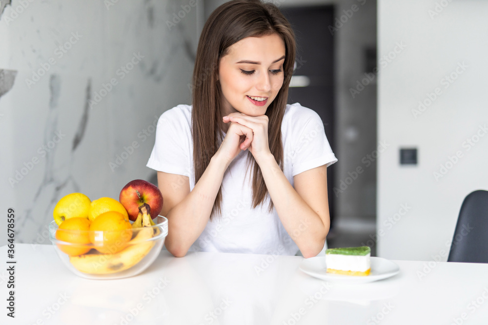 Young smiling woman with green apple in one hand choosing between healthy and unhealthy food in the kitchen. Difficult choice between fresh fruit vegetables or sweets. Dieting. Diet. Healthy Food Young smiling woman with green apple in one hand choosing between healthy and unhealthy food in the kitchen. Difficult choice between fresh fruit vegetables or sweets. Dieting. Diet. Healthy Food
