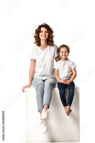 happy mother and daughter looking at camera while sitting on cube on white