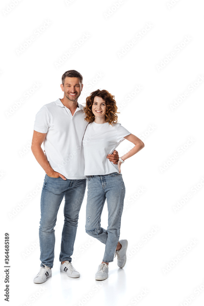 cheerful man standing with hand in pocket near wife with hand on hip on white