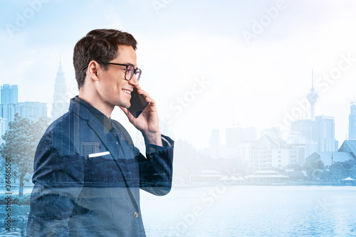 Prosperous handsome entrepreneur in suit and glasses pensively talking phone and looking on Kuala Lumpur cityscape. The concept of problem solving consulting . KL skyscrapers. Double exposure. photo