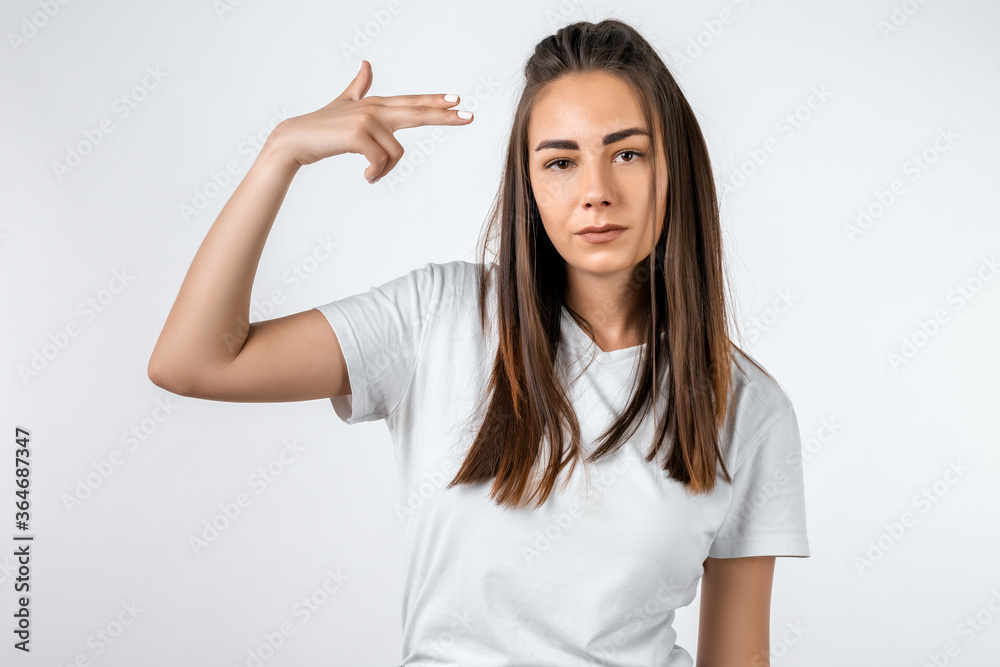 I'm fed up with this! Stylish European girl with long chestnut hair shoots in temple, tilts head, dressed in casual t shirt, demonstrates suicide gesture, isolated on white background I'm fed up with this! Stylish European girl with long chestnut hair shoots in temple, tilts head, dressed in casual t shirt, demonstrates suicide gesture, isolated on white background