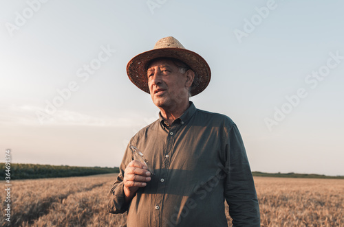 Portrait of senior farmer with hat standing in wheat field at sunset.