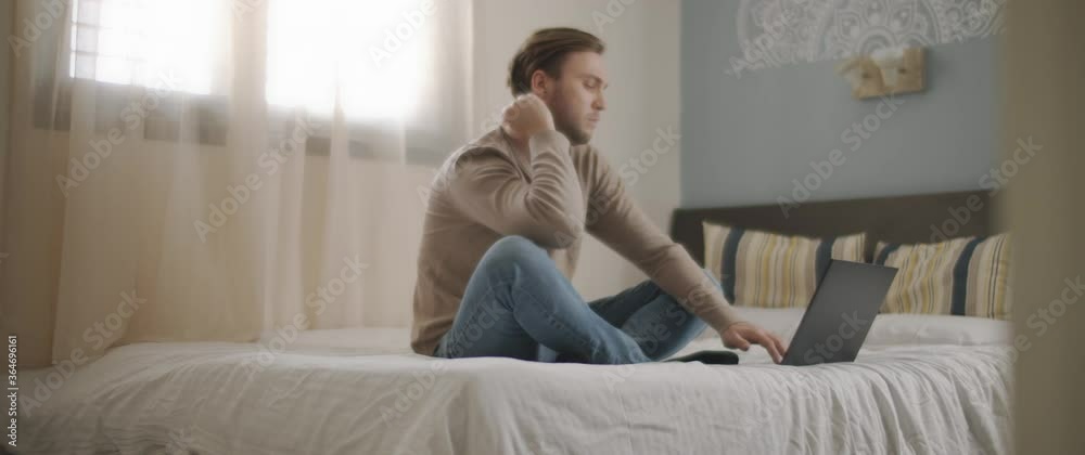 A young man sitting in bed with laptop, working from home. Slow motion, medium shot, BMPCC 4K A young man sitting in bed with laptop, working from home. Slow motion, medium shot, BMPCC 4K