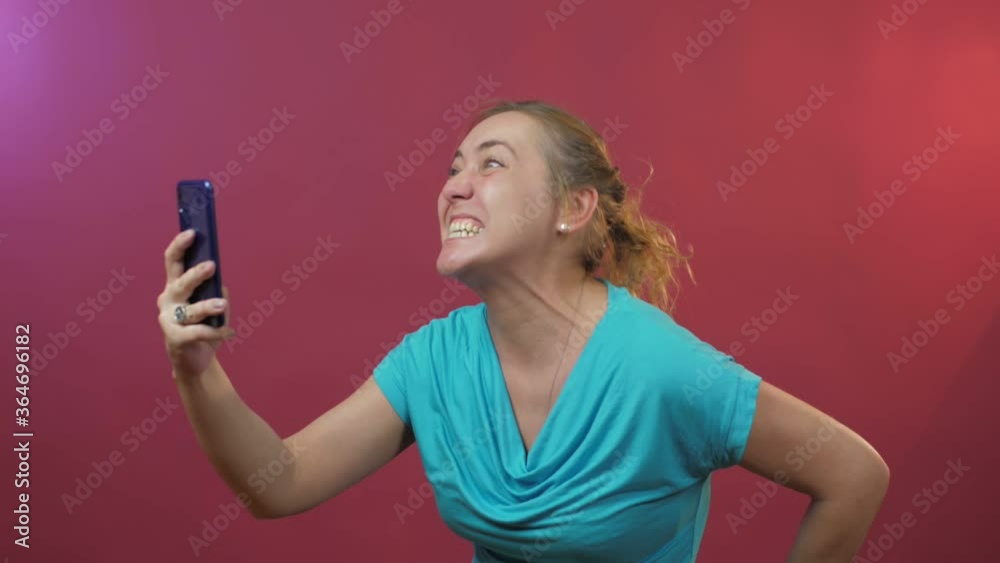 joyful young woman with fair hair makes selfie on black smartphone pulling faces against pink background close view joyful young woman with fair hair makes selfie on black smartphone pulling faces against pink background close view