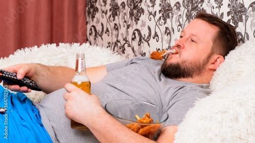 fat man drinks beer and eats unhealthy food chicken wings, bored in front of the TV outlook on the couch. The concept of malnutrition, quarantine at home, alcoholism.