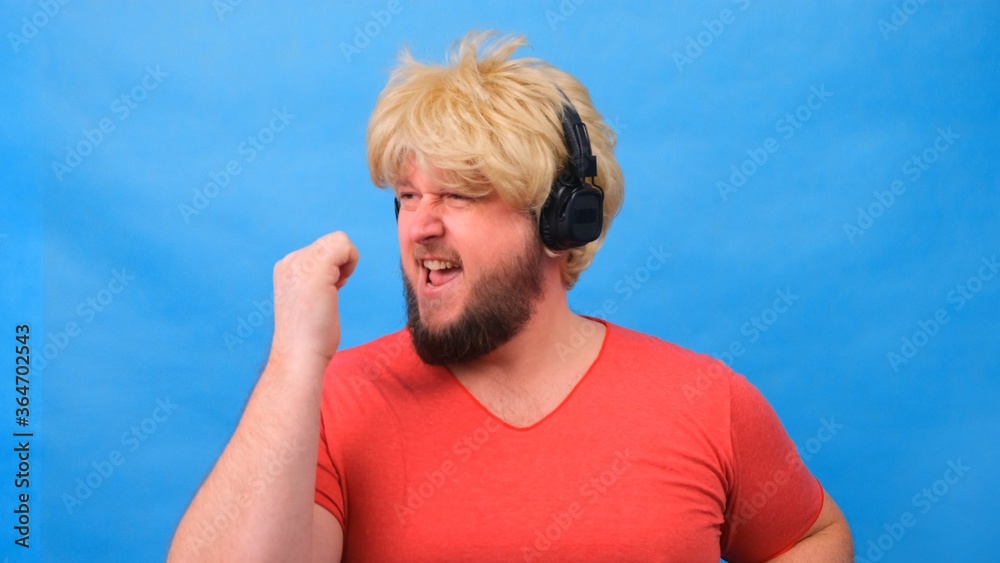 Naklejka premium freaky fat man in a wig and a pink T-shirt in his hands listens to music on headphones and dances on a blue background