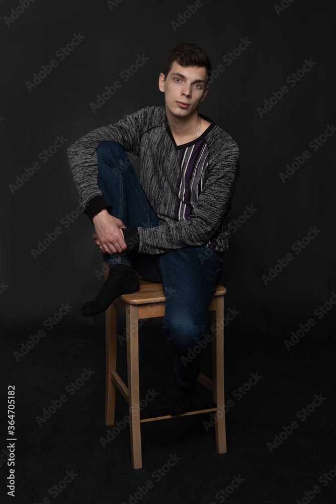 Portrait of serious young man is sitting on chair with one leg on it on black background