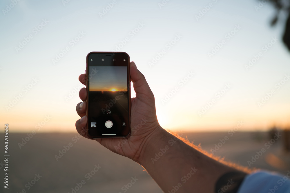 Fototapeta premium Cropped image of man's hand holding modern smartphone using application for taking evening photos of beautiful sundown, closeup view of male shooting video of sunset and skyline on telephone camera