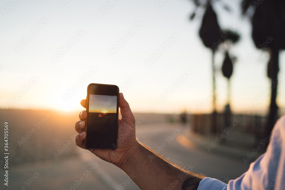 Fototapeta premium Close up view of male shooting video of sunset and skyline on smartphone camera, cropped image of man's hand holding mobile phone using app for making pictures of sundown during summer vacation