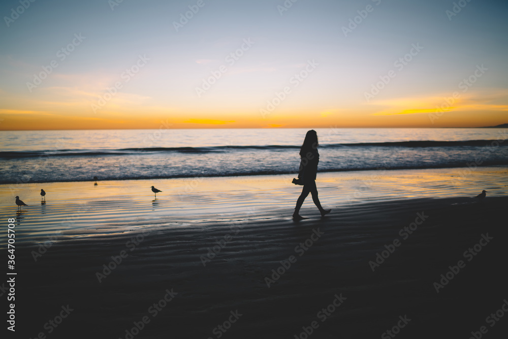 Lonely woman silhouette on sandy shore of tranquil ocean during sceni sunset, upset girl walking near sea on coastline feeling depressed recreating with beautiful nature landscape in evening.
