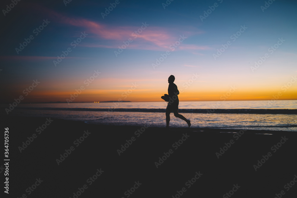 Young male person silhouette running on sandy ocean shore in evening twilights after sunset, guy jogging spending active summer weekends on sea coastline enjoying scenic view of sky and dawn.