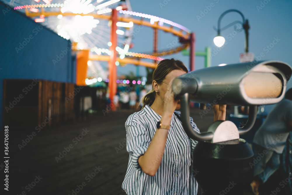 Hipster girl looking at view in coin operated optical binocular standing in amusement park shining with night lights recreating on summer vacations, female tourist resting in Santa monica pier.