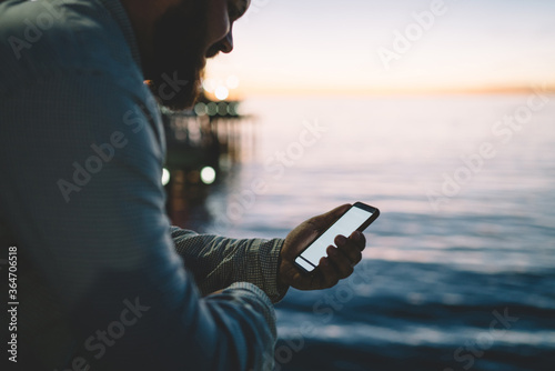 Cropped image of positive man smiling while reading message on mobile phone with mock up screen standing outdoors, smiling guy satisfied with 4G internet connection in roaming for online chatting.